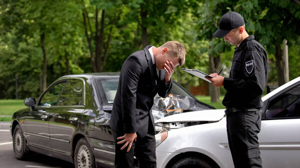 Desperate businessman covering face, policeman writing accident report collision