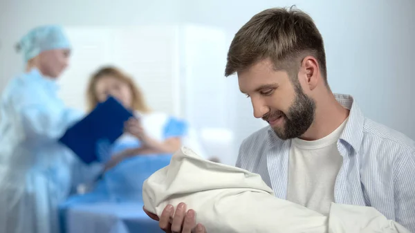 出産後のケアを妻に説明する看護師の間に新生児を抱く幸せな父親 — ストック写真