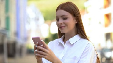 Neşeli beyaz kız dışarıda akıllı telefondan mesaj atıyor ve gülümsüyor.