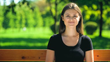Parkta bankta oturan ve gülümseyen mutlu genç bir kadın. Hafta sonu boş zamanlarında dışarıda.