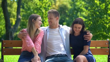 Handsome man resting on bench in park hugging two attractive females, ladies man clipart