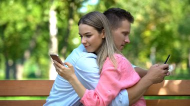 Parkta kucaklaşan genç çift akıllı telefonlarda sohbet ediyor, iletişim eksikliği...