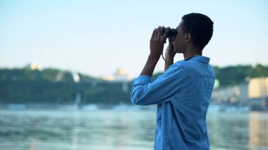 Nehirdeki teknelere dürbünle bakan melez bir çocuk. Gençlik rüyaları.