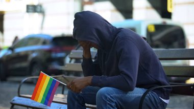 Kukuletalı siyahi adam, elinde lgbt azınlık bayrağı, önyargıları ile bankta oturuyor.