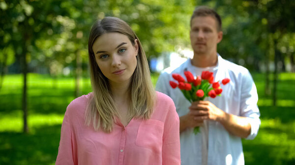 Annoyed young female looking camera, loving young man with flower on background
