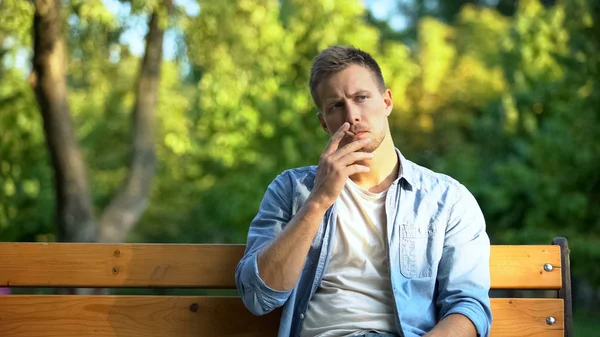 Pensive young man smoking cigarette relaxing on bench in park, harmful habit