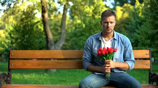 年轻男子与红色郁金香坐在公园的长椅上等待女朋友的约会 — 图库照片