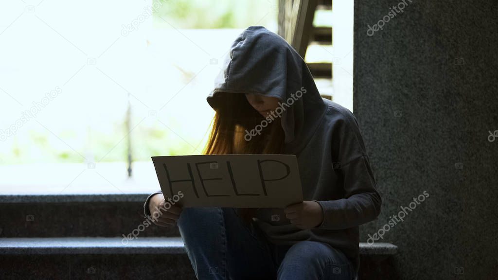Homeless teenage girl in hoodie showing help sign on stairs, social insecurity