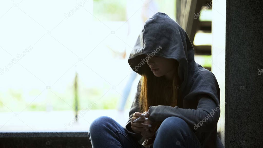 Offended teenage girl in hoodie secretly smoking cigarette on stairs, depression