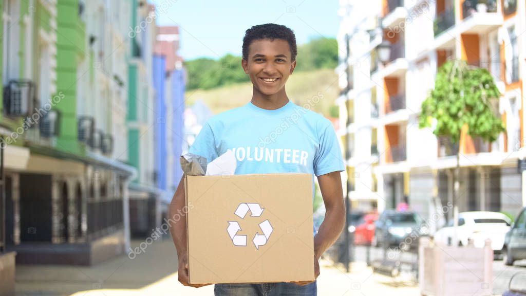 Happy mixed-race teen male eco-volunteer holding paper box with recycling sign