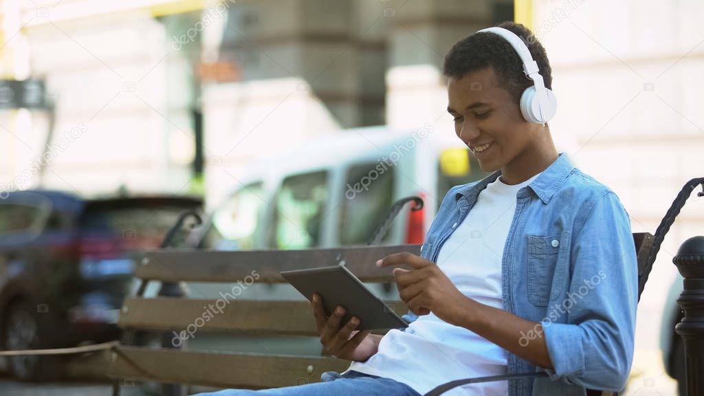 Cheerful mixed-race boy in headphones using tablet outdoors, listening to music