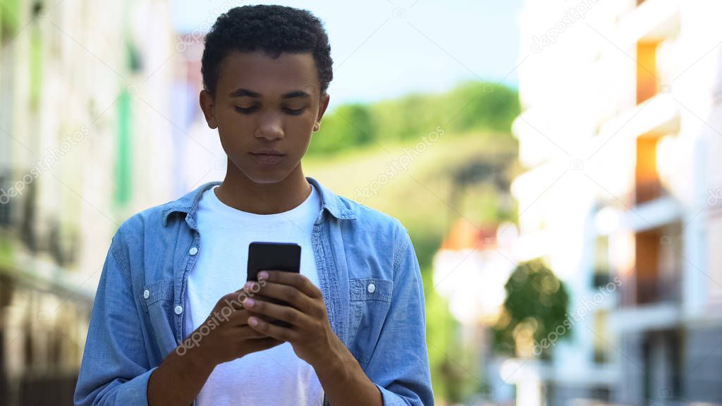 Afro-American male teenager chatting smartphone outdoors, online communication