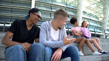 İki erkek genç yanında oturan kadın sınıf arkadaşlarına gülüyor, gençlik dedikoduları