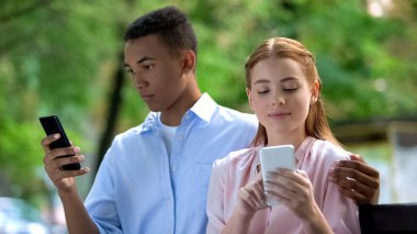 Parkta bankta oturan çok ırklı genç çift sarılma, sohbet akıllı telefon