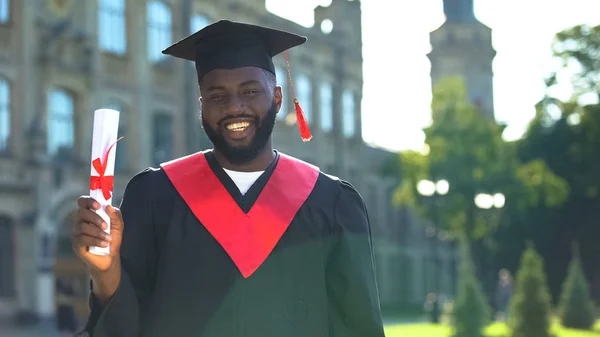 Excited afro-american student in graduation gown holding diploma looking camera