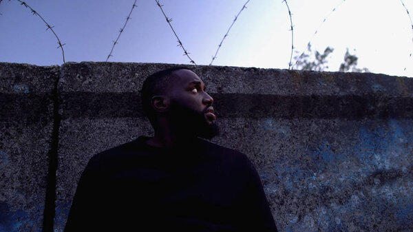 Заключенный афро-американец смотрит на колючую проволоку, лагерь беженцев, безнадежность
