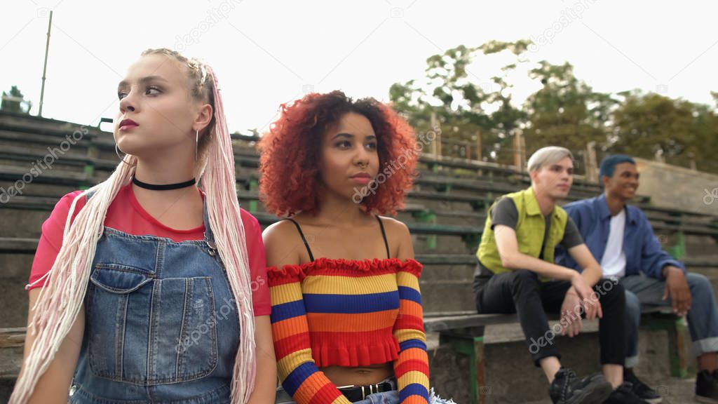 Stylish teenage girls sitting on stadium bench apart from boys, feeling shy