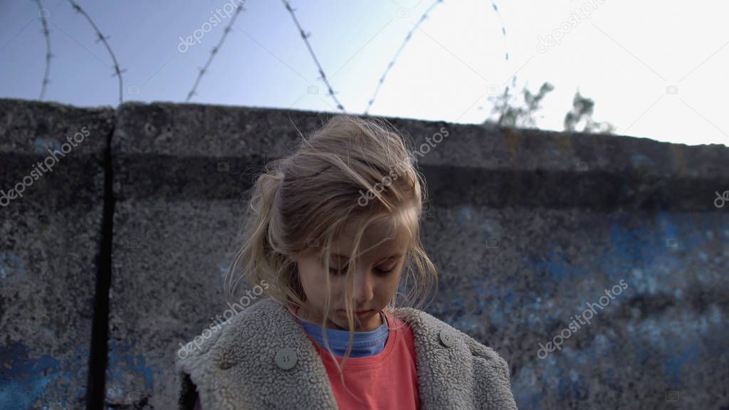 Little girl with muddy face standing at barbed wire, war child in refugee camp