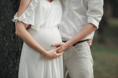 Beyaz açık yaz elbiseli hamile gelin ve parlak elbiseli damat, büyük hamile karnına elleriyle sarılıyorlar, yüzleri görünmüyor.