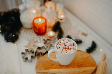 Noel kartı, beyaz bardakta sıcak bir içecek, beyaz marşmelovlar ve ahşap tezgahın üstünde kırmızı tepsi, yanan mumlar ve çelenklerle dolu beyaz bir masa.