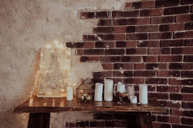 Duvarın yarısı tuğla ve gri macun, Noel süsü beyaz mumlar, dekorasyonlar ve ahşap bir masada Noel 'le ilgili sözlerin olduğu bir resim.