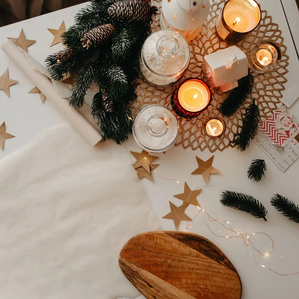 Noel Masası Dekoru, bir rulo kağıt, yanan beyaz ve kırmızı mumlar, ahşap kesme tahtası, Noel ağacı dalları ve kağıt yıldızlar, beyaz bir masanın üzerinde, kare şeklinde.