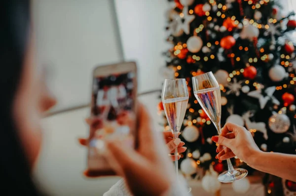 Kız beyaz bir telefonla fotoğraf çekiyor, iki kadeh şampanya ile birlikte bayanların elindeki Noel ağacının arka planında.