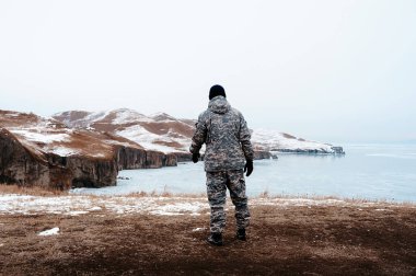 Askeri giysili bir adam kışın arka planında kamuflaj yapıyor ve bembeyaz bir kar yağışı var. Bir avcı inanılmaz kuzey manzarasına geri çekiliyor. Buz denizi ve buzdaki tepeler.