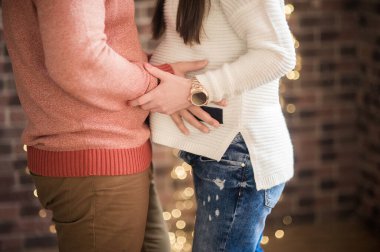 Yüzü olmayan bir çift aşık karşı karşıya durur ve beyaz, kalın kazaklı hamile bir kızın karnına sarılır tuğla bir duvara ve Bokeh 'li çelenklere karşı.