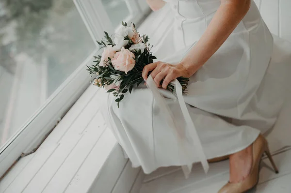 控えめなシンプルな白いドレスを着たスタイリッシュな花嫁は 牡丹のバラと装飾のための緑の植物からピンクのほこりの多い花の信じられないほどの花束を保持しています — ストック写真