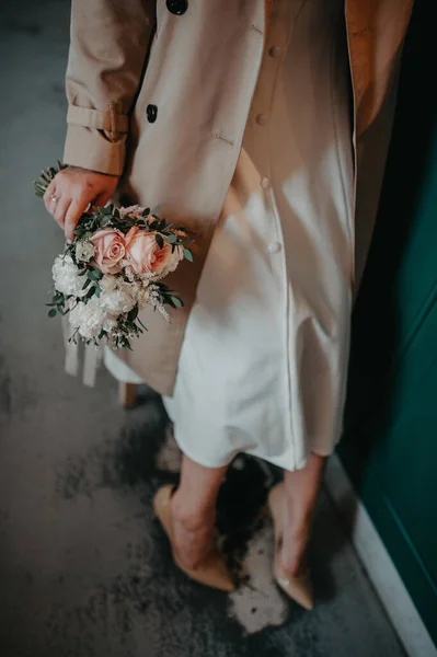 膝までエレガントな白いドレスに身を包んだ超スタイリッシュな花嫁の横のイメージと ほこりっぽいピンクのバラの花束を持つベージュのトレンチコート — ストック写真