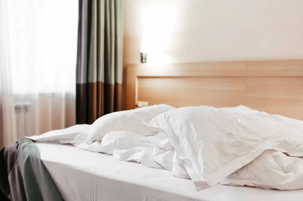 crumpled white factory bedding on the bed with a blanket and pillows, next to the bedside table and dark heavy curtains on the floor, a standard elegant look of a hotel room