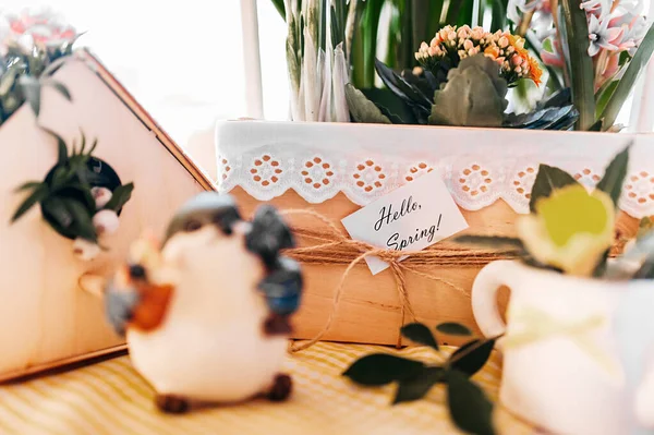 spring decor, a wooden vase with green flowers, a moth and the inscription hello spring, on a wooden table on which there is a blue bird, a wooden decorative birdhouse with flowers and a white porce