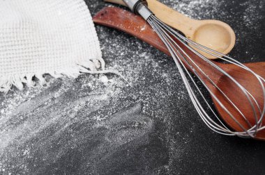 Çevrimiçi yemek yapma planı. Siyah kaplama dökülmüş un, metal çırpı ve tahta kaşıklar beyaz pamuklu mutfak havlusunda. Yukarıdan manzara. Metin için boşluk