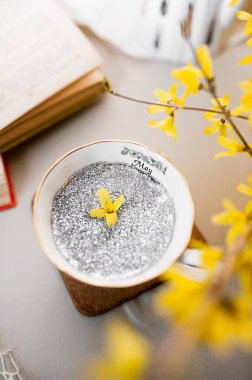 Metni gümüş pullarla dolu beyaz porselen bardak ve çiçek açan sarı çiçek buketi ve eski kitaplar karşısında küçük sarı bir çiçek. en üstteki görünüm