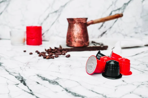 Red and black coffee capsules on the background of a Metal Turkish coffee pot with a long wooden handle and scattered coffee beans fried on white marble