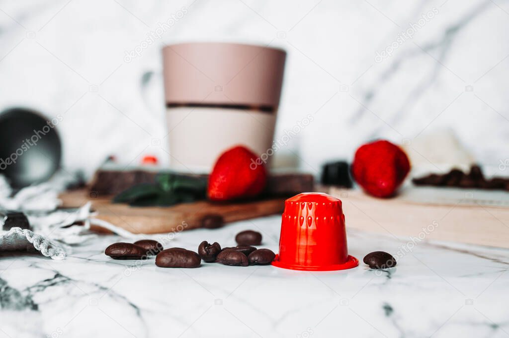Red coffee capsules on the background of dusty pink pastel mug and scattered coffee beans fried on white marble