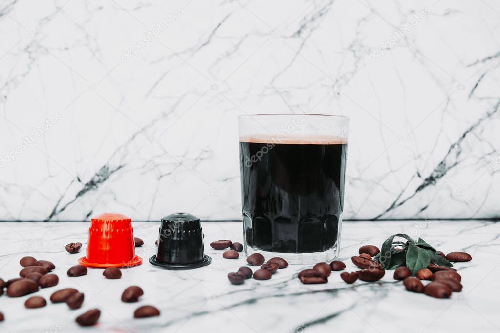 Red and black coffee capsules and clear glass mug with strong black coffee in scattered grains on white marble.
