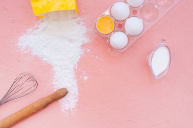 Beyaz yumurtalar, ahşap bir oklava, metal bir çırpı ve pembe arka planda sarı bir torbadan saçılmış un. Pişirme için mutfak gereçleri, hamur hazırlama