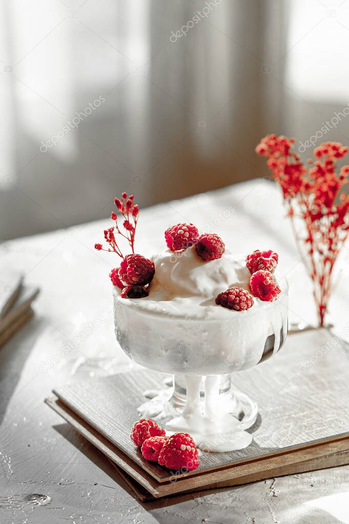 bowl with white ice cream, red berry jam, fresh raspberries decorated with red flowers on light table in the sunset light