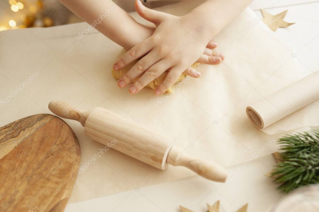 children hands crumple dough on baking paper on a light table, Christmas tree branches, a small rolling pin, a wooden cutting Board and blurred bokeh lights from a garland on the background