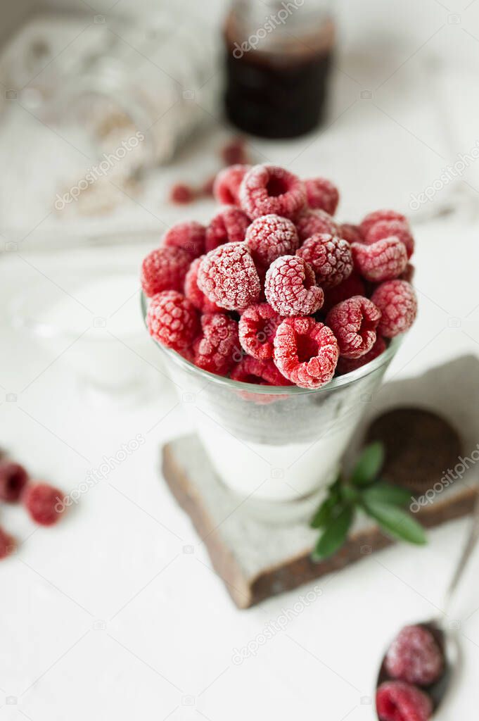 healthy and beautiful summer cooling food. Fresh cold raspberries, layer of chocolate chips and yogurt in glass on light table with scattered berries, mint leaves, cans of muesli and jam on background