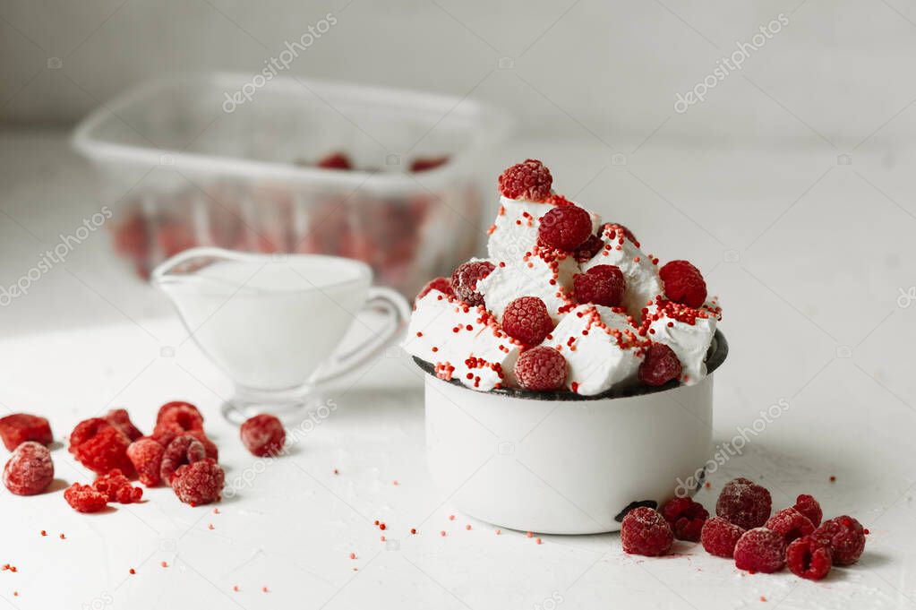 Slices of white milk ice cream with fresh red berries of ripe raspberries and red sprinkles in a white metal mug on a light table, a bowl of milk and a bowl of berries are blurred on the background