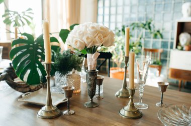 Beyaz bir buket taze, tıpatıp aynı güller ahşap bir masa üzerinde metal gümüş çatal bıçak, şamdanlar ve eski mumlarla. Klasik tarzda düğün dekoru..
