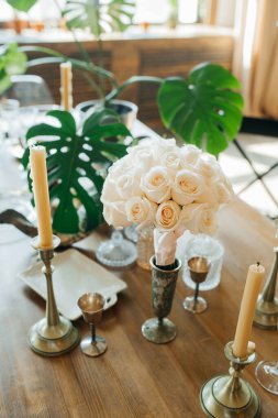 Beyaz bir buket taze, tıpatıp aynı güller ahşap bir masa üzerinde metal gümüş çatal bıçak, şamdanlar ve eski mumlarla. Klasik tarzda düğün dekoru. Dikey