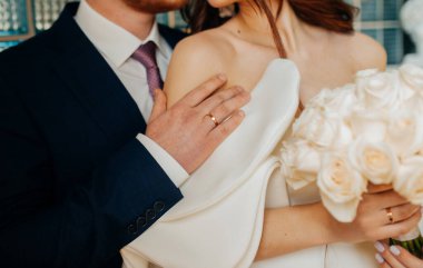 Şık bir düğün fotoğrafı. Mavi takım elbiseli damat, gelini yumuşak kumaştan yapılmış beyaz bir elbiseyle nazikçe kucaklıyor. Kabarık gelinlik kollu, fırfırlı kollu olağandışı bir gelinlik..