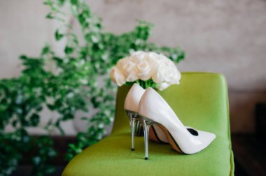 Uzun şeffaf topuklu sivri uçlu beyaz bir düğün ayakkabısı açık yeşil bir sandalyede gri beton bir duvara ve ev yeşili bir çiçeğe karşı bir düğün buketi. Gelin için aksesuarlar.