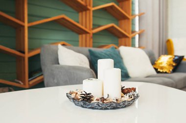 Aromaterapi için şamdandaki beyaz mumlar gri kanepeli, rahat yastıklı ve kitaplar için ahşap raflı rahat bir dairenin arka planına karşı.