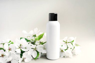 Beyaz arka planda yeşil yapraklı, yemyeşil çiçek filizleri açan siyah şapkalı beyaz bir şişe. Kozmetik çizimi. Cilt ve vücut bakımı eşleşir