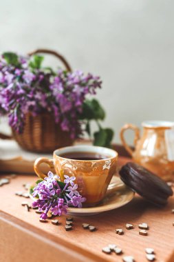 Yeşillik taze çiçeklerle ev yapımı çay partisi. Siyah çayla altın porselen fincan, çikolatalı pasta, mor leylak çiçekleri hasır sepetteki ahşap tepside kalplerle süslenmiş